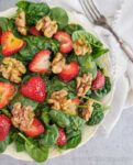 Spinach salad with sliced strawberries and walnuts in a white bowl with a napkin and fork.
