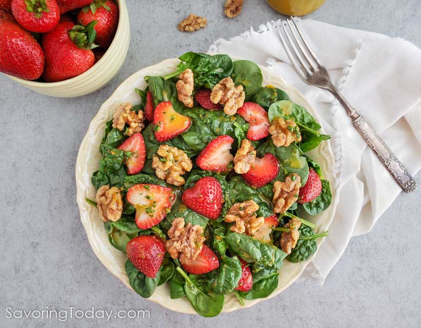 Spinach salad with strawberries and walnuts on a cream plate with a bowl of strawberries on the side. 