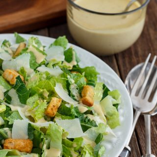 Close-up of Caesar Salad in a white bowl with salad tongs and dressing beside it.