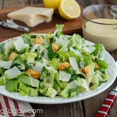 Romaine lettuce dressed with Caesar dressing in a white bowl over a red striped towel with parmesan and lemon on a cutting board in the background. A cup of the dressing in beside the bowl.