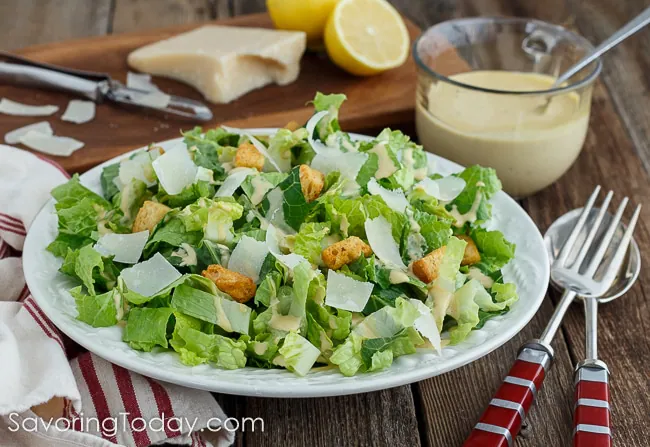 Romaine lettuce dressed with creamy Caesar dressing in a white bowl over a red striped towel with Parmesan and lemon on a cutting board in the background. 