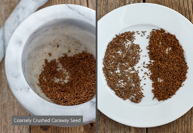 Crushed caraway seeds in a mortar and pestle beside the seeds on a white plate.