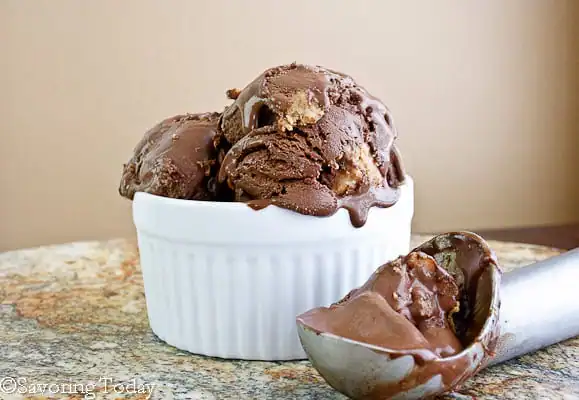 Chocolate ice cream with peanut butter chunks in a white ramekin with the ice cream scoop beside it.