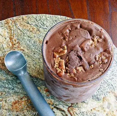 Chocolate ice cream with peanut butter chunks in a white ramekin with the ice cream scoop beside it.