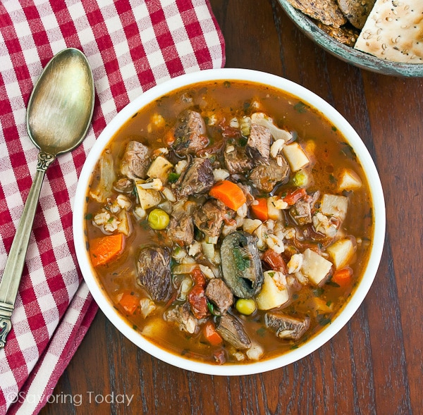 Roast Beef and Vegetable soup showing chunks of beef, mushrooms, carrots, peas, potatoes and beef broth. Served in white bowl with a red and white napkin, seeded crackers and spoon.