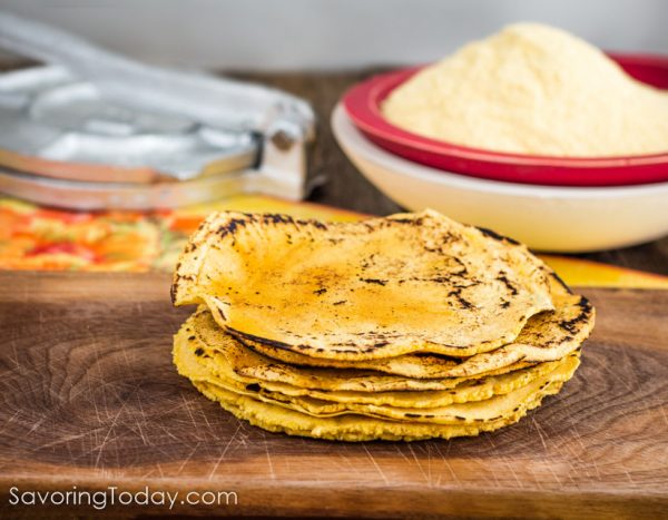 Homemade Corn Tortillas stacked on a cutting board with masa flour and tortilla press.