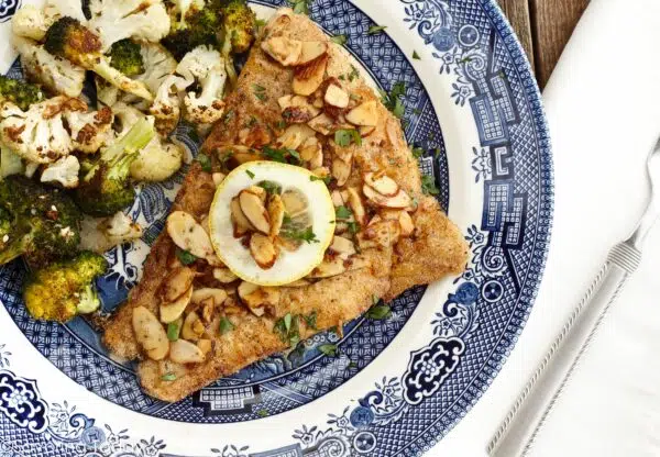 Pan-seared fish with toasted sliced almonds and a slice of lemon served on a blue and white plate beside a fork and white napkin.
