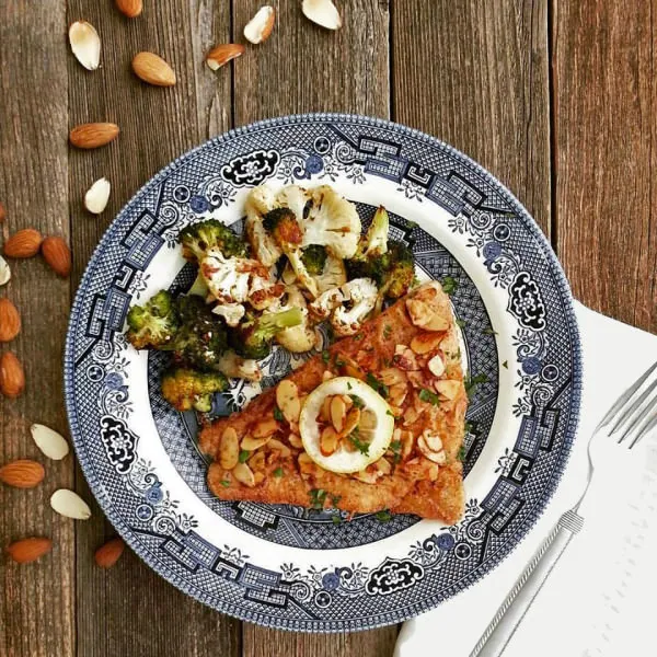 Pan-seared fish with sliced almonds and a slice of lemon with a side of roasted vegetables on a blue and white plate. 