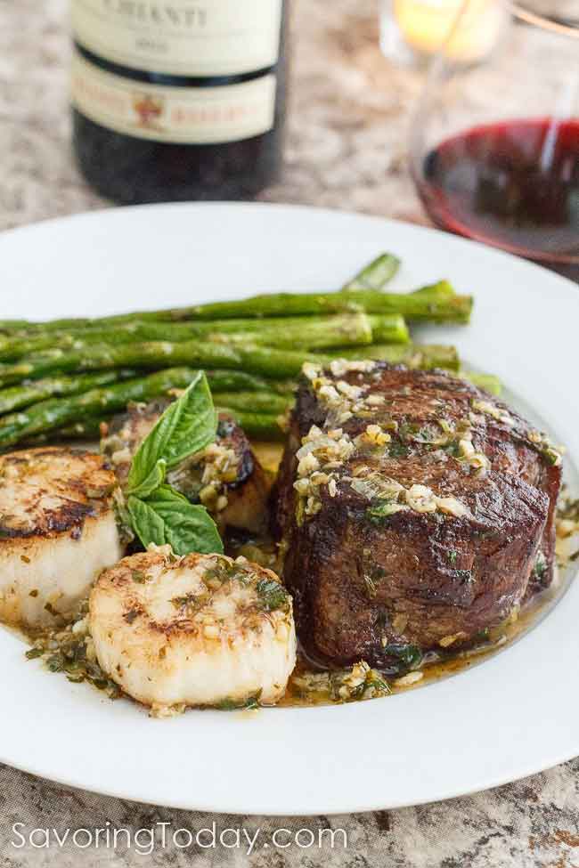 Pan seared steak and scallops with a garnish of basil leaf and asparagus served on a white dinner plate.