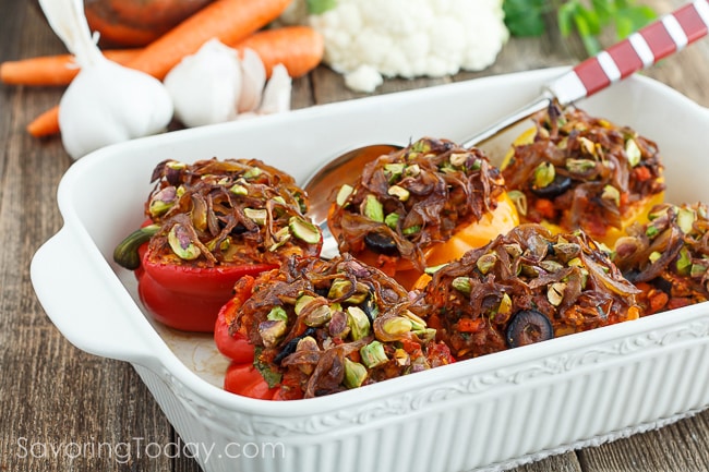 Red and orange bell peppers stuffed with sausage, cauliflower, and black olives, topped with caramelized onions and pistachios. Peppers are arranged in a white ceramic baking dish and serving spoon with garlic, carrots and cauliflower in the background.