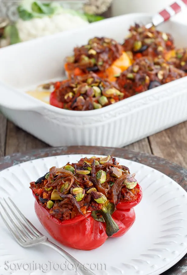 A stuffed red bell pepper topped with caramelized onions and pistachios on a white plate with a fork. Pan of stuffed peppers in the background. 