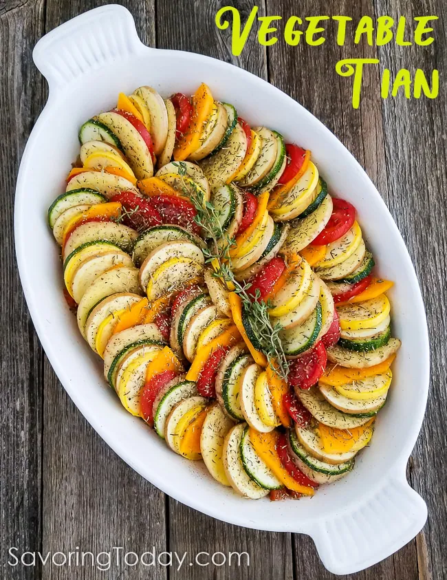 Alternating vegetables-tomato, squash, potato-in a white baking dish with dried herbs sprinkled on top.