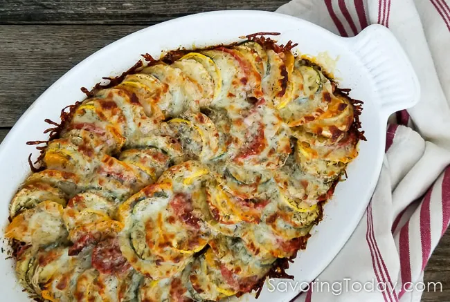 A baked vegetable gratin in a white baking dish beside a red striped towel on a wood table.