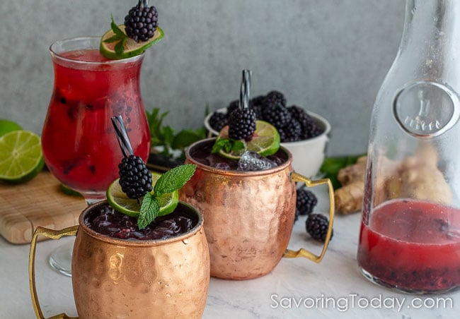 Cocktail in copper mugs and clear glass beside a liter pitcher and scattered blackberries.