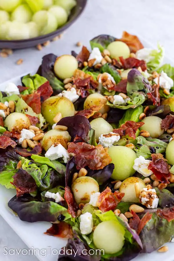 Salad with melon balls, goat cheese, prosciutto and pine nuts on a white plate on marble counter.