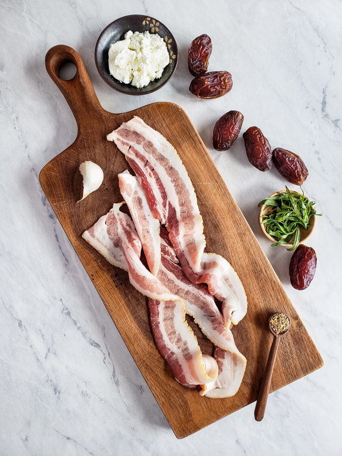 Bacon and clove of garlic on a cutting board with goat cheese, dates, and fresh tarragon beside the board.