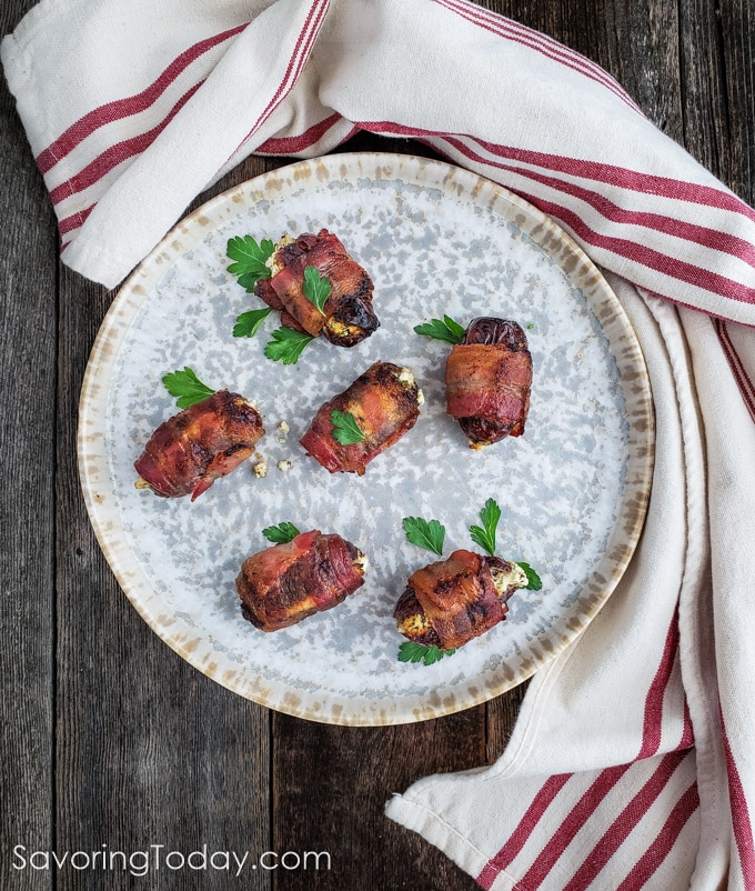 Bacon-wrapped dates stuffed with herbed goat cheese on a grey plate with a red striped towel around it.