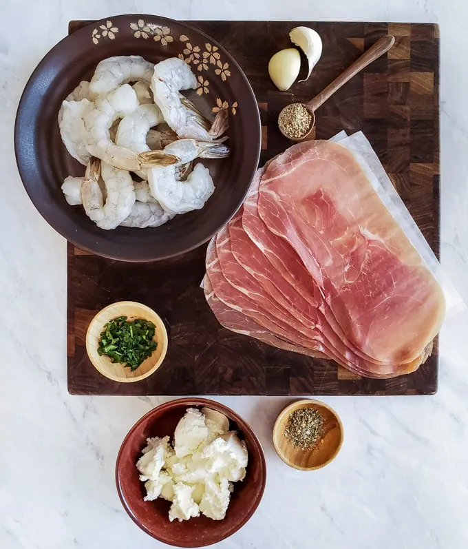 Prosciutto, shrimp, goat cheese, parsley, lemon pepper, garlic, and Italian herbs on a cutting board to show the ingredients in a shrimp appetizer recipe.