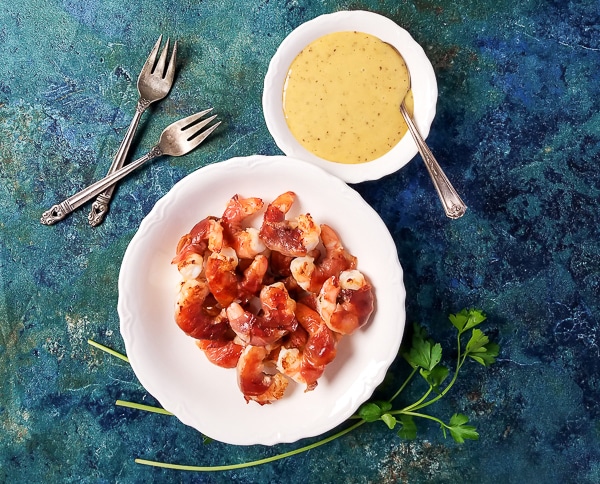 Sweet-tangy honey mustard sauce beside prosciutto-wrapped shrimp in a white bowl.