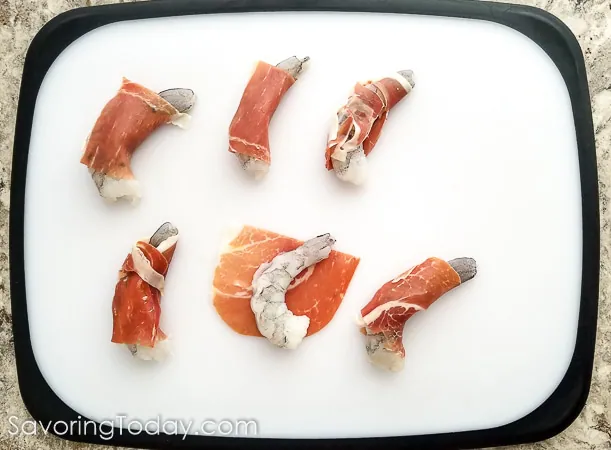 Six shrimp wrapped in prosciutto on a cutting board.