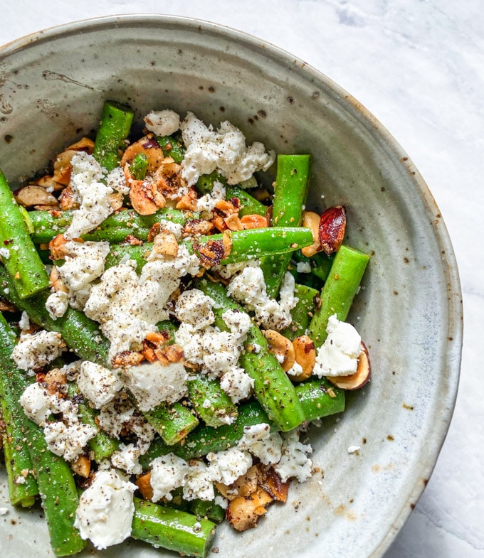 Brown butter and hazelnuts coat green beans in a dish with goat cheese sprinkled on top.