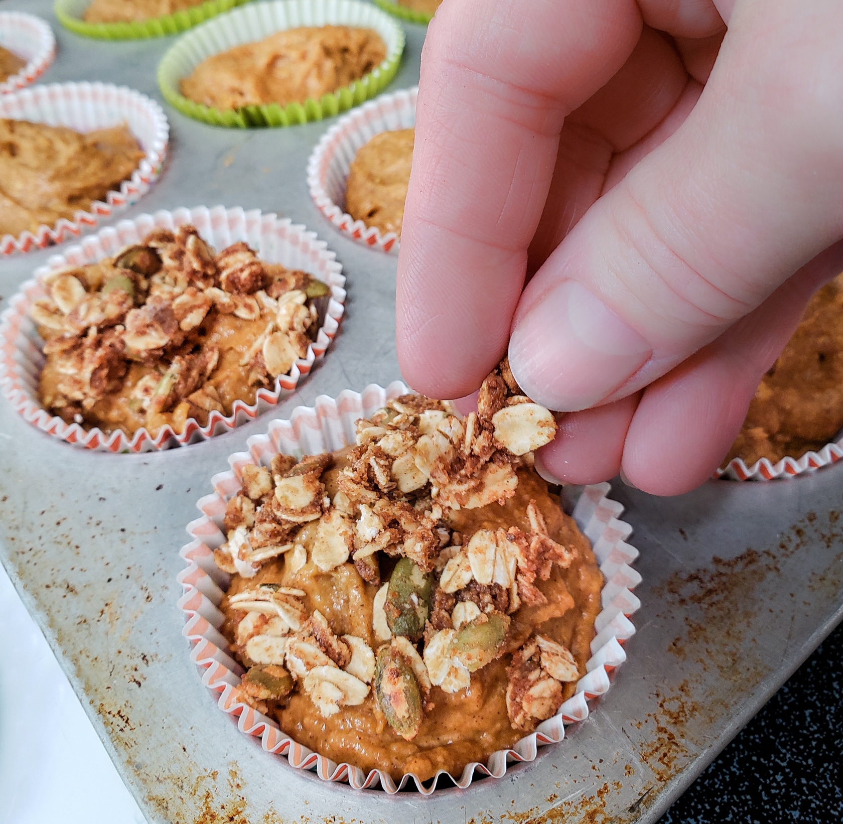 Fingers sprinkling crumb topping onto unbaked pumpkin muffins.
