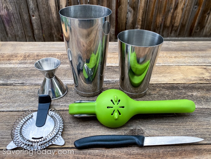 A shaker tin, strainer, juicer, knife, and jigger for bartending.