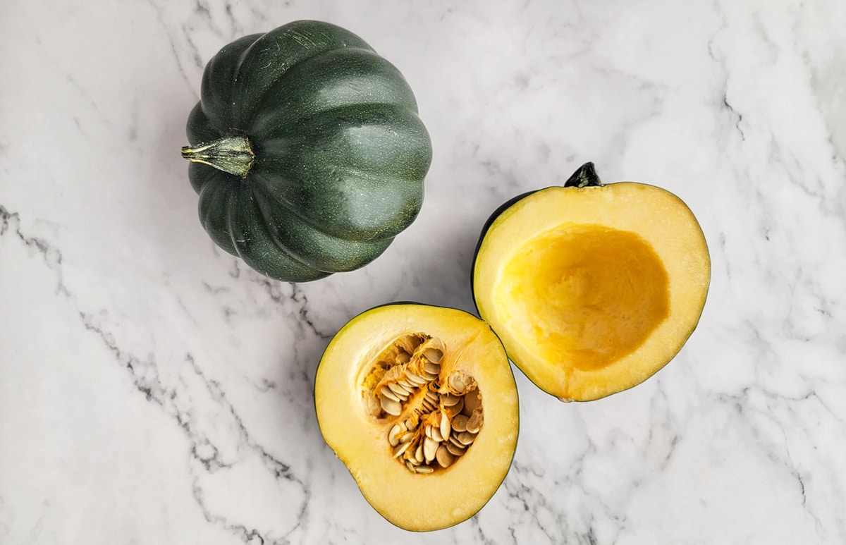 Ripe acorn squash whole and one sliced in half with the seeds scooped out of one on a counter.