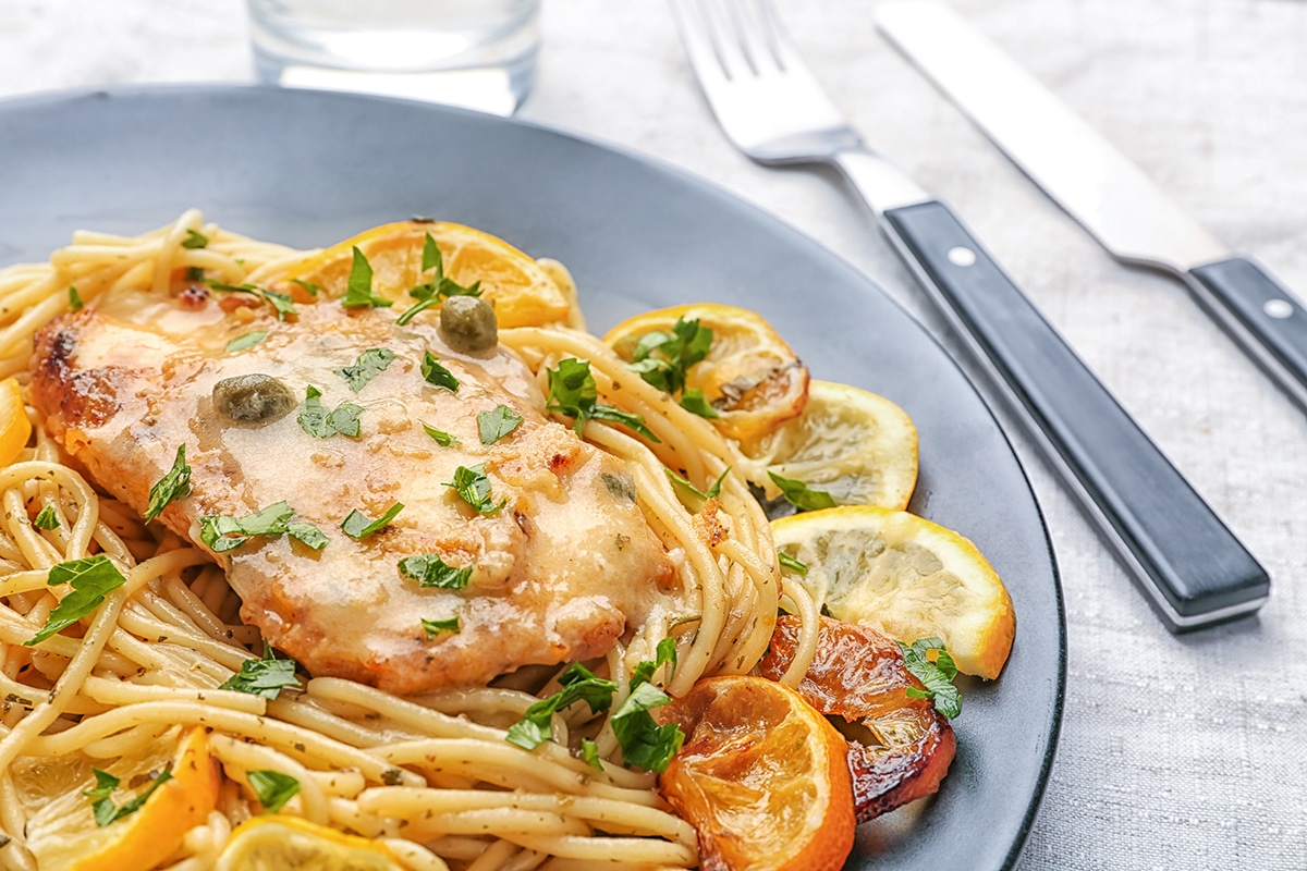 Chicken piccata served with pasta on a black plate with knife and fork