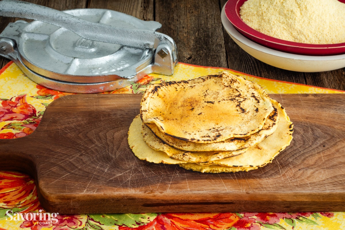 stacked corn tortillas on a wood cutting board with a press and masa harina in the background