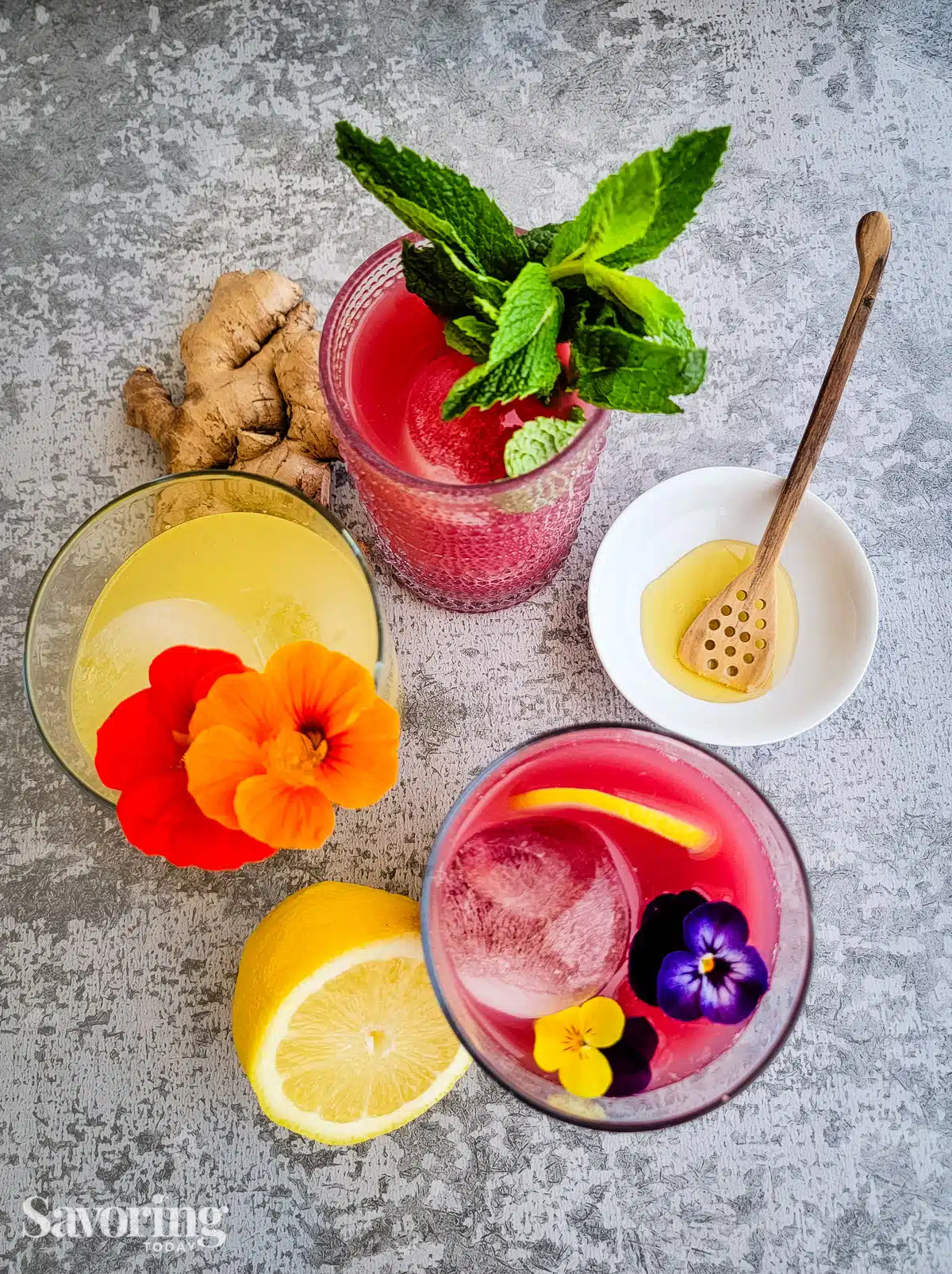 drinks on a counter garnished with flowers and mint beside fresh ginger and lemon