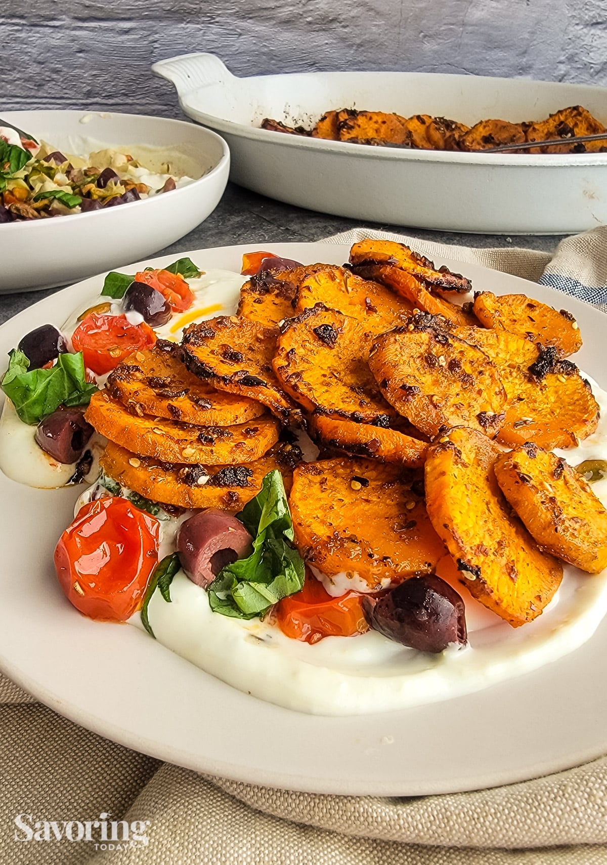 roasted sweet potates on  whipped ricotta with tomatoes and olives on a tan plate beside casserole dishes