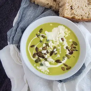 Asparagus soup with pepitas and feta cheese in a white bowl with bread